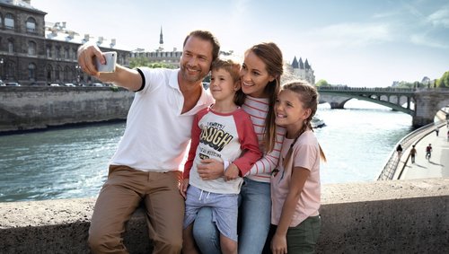 Familie mit Vater, Mutter, Sohn und Tochter macht Selfie auf Brücke über Fluss in Paris bei sonnigem Wetter.