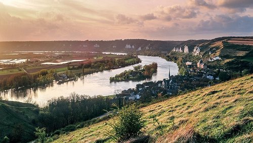 Fluss Seine, der sich durch grüne Hügel und das Dorf Les Andelys mit Kirche und Felsenlandschaft schlängelt bei Sonnenuntergang.