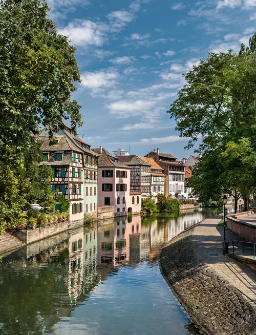 Ein ruhiger Kanal in Straßburg, gesäumt von Fachwerkhäusern und Bäumen, spiegelt sich im Wasser wider. Ein Gehweg auf der anderen Seite des Kanals.