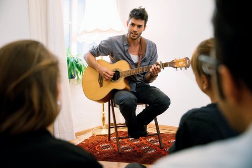 Der Musiker Henrik Wiards spielt akustische Gitarre bei einem intimen Konzert. Er sitzt auf einem Stuhl ihm gegenüber eine kleine Gruppe Zuhörer in einem Wohnzimmer mit Teppich und Pflanzen.