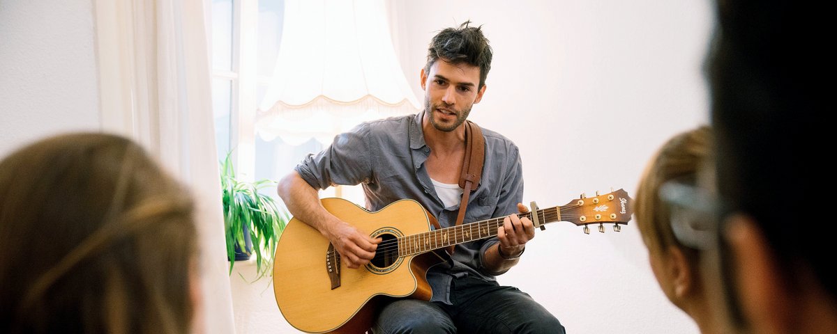 Der Musiker Henrik Wiards spielt akustische Gitarre bei einem intimen Konzert. Er sitzt auf einem Stuhl ihm gegenüber eine kleine Gruppe Zuhörer in einem Wohnzimmer mit Teppich und Pflanzen.