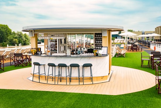 Das Sonnendeck der A-ROSA SENA bei Abenddämmerung mit stimmungsvoll belechteter UP & DOWN BAR und Sitzgruppen, Blick auf den Fluss und das beleuchtete Amsterdam.