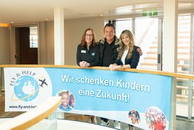Drei Personen stehen hinter einem blauen Banner mit der Aufschrift 'Wir schenken Kindern eine Zukunft' und dem Logo von Fly & Help in einem hellen Innenraum