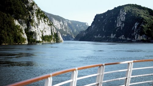 Blick von einem Flusskreuzfahrtschiff auf die bewachsene Felsenlandschaft im Donaudelta. Im Vordergrund ist eine Reling und dahinter der stille Fluss.
