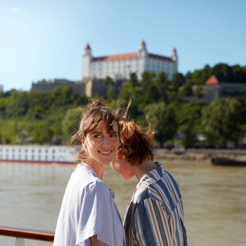 Zwei Frauen stehen an der Reling auf einem Flusskreuzfahrtschiff auf der Donau mit Blick auf ein weiteres Schiff und der Burg Bratislava auf einem bewachsenene Hügel im Hintergrund.