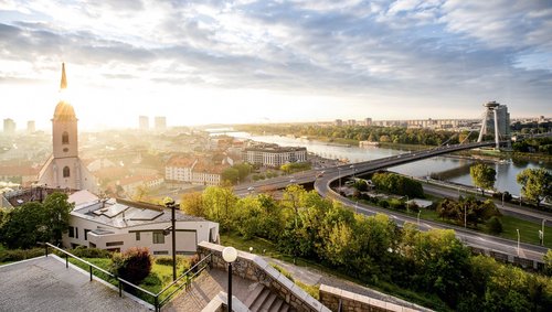 Blick von einer Anhöhe auf Bratislava mit Burg, Brücke und der Donau im Vordergrund bei Sonnenaufgang