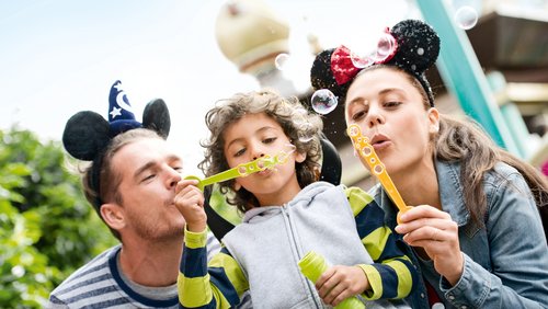 Zwei Kinder und ihr Vater im Disneyland Paris. Der Vater und das Mädchen rechts haben Micky-Maus-Ohren auf dem Kopf. Der Junge in der Mitte und das Mädchen pusten Seifenblasen.