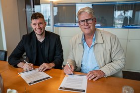 Zwei Männer sitzen an einem Tisch und unterschreiben Verträge, rechts Rolf-Dieter Maltzahn trägt hellblaue Hemd und helle Jacke