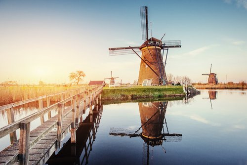 Traditionelle niederländische Windmühlen in einer grünen Landschaft mit blauem Himmel am Kanal von Rotterdam. Im Vordergrund eine schmale Holzbrücke über dem Kanal.